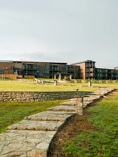 The image shows a large modern building complex surrounded by open grass fields and a stone pathway, set under a clear sky.