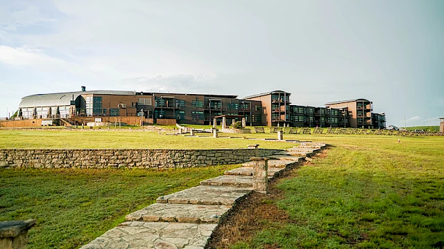 The image shows a large modern building complex surrounded by open grass fields and a stone pathway, set under a clear sky.