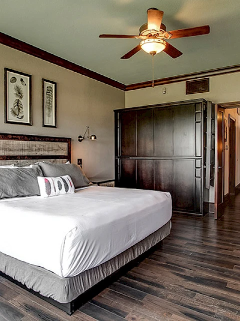 The image shows a hotel room with a large bed, armchair, ceiling fan, wooden floor, and wall art, creating a cozy atmosphere.