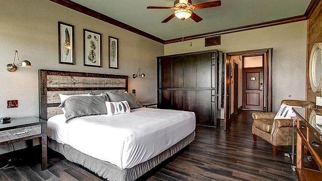 The image shows a hotel room with a large bed, armchair, ceiling fan, wooden floor, and wall art, creating a cozy atmosphere.