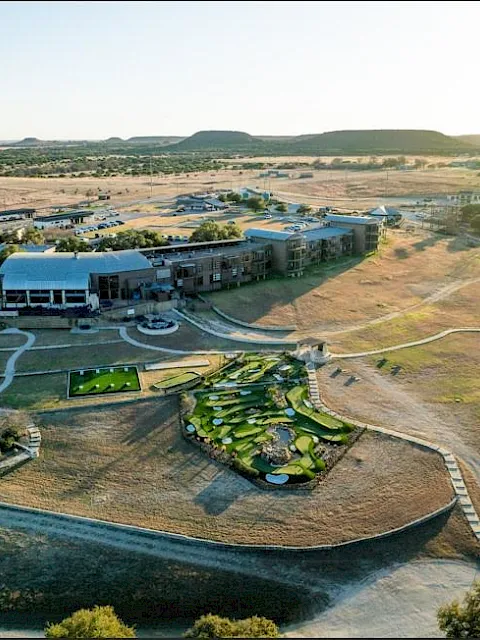Aerial view of a resort complex with hotel buildings, pools, gardens, and winding paths amid dry fields and hills.