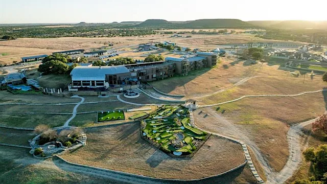 Aerial view of a resort complex with hotel buildings, pools, gardens, and winding paths amid dry fields and hills.