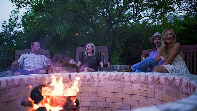 Four people sit around a stone fire pit with a burning fire, relaxing and enjoying each other's company outdoors.
