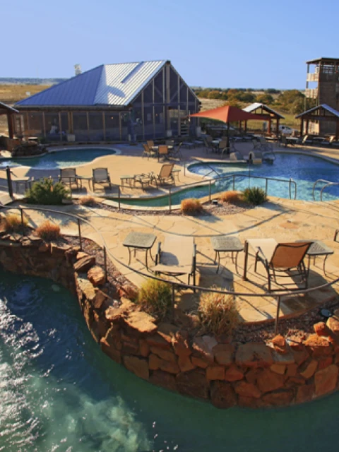 Outdoor leisure area with a pool, chairs, and tables surrounded by a rocky landscape under a clear blue sky.