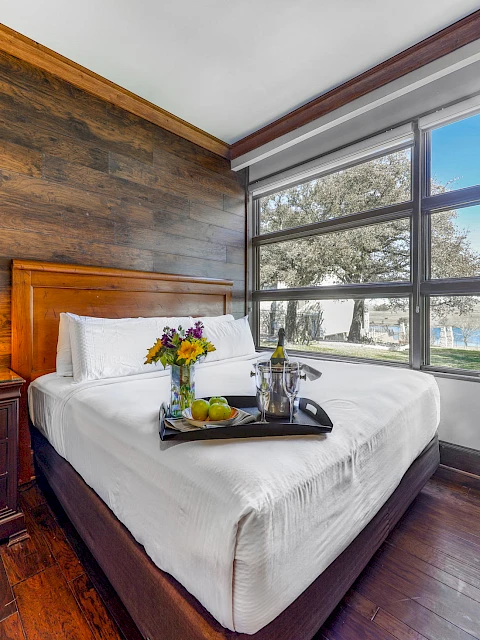 The image shows a cozy bedroom with a wooden accent wall, large window view, bed with a tray of flowers, wine, fruit, a lamp, and a leather chair.