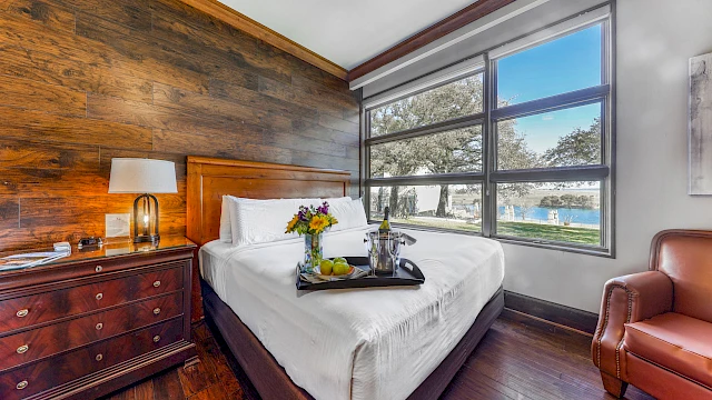 The image shows a cozy bedroom with a wooden accent wall, large window view, bed with a tray of flowers, wine, fruit, a lamp, and a leather chair.