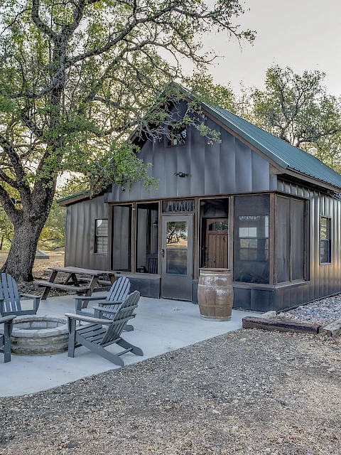 A small cabin with a metal exterior, surrounded by trees, has outdoor seating around a fire pit on a concrete slab.