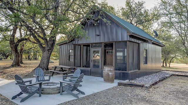 A small cabin with a metal exterior, surrounded by trees, has outdoor seating around a fire pit on a concrete slab.