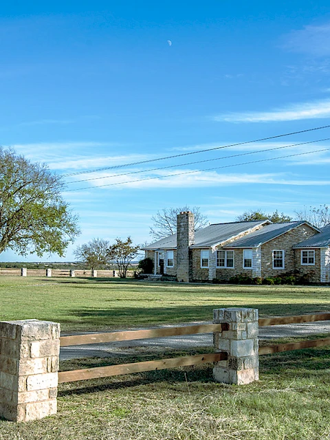 A stone house with a chimney is surrounded by trees, a green lawn, and a wooden fence under a clear blue sky.