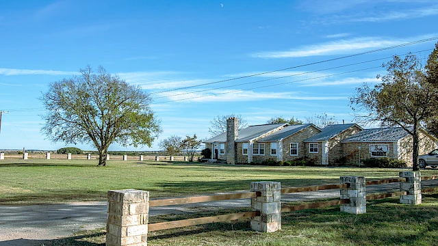 A stone house with a chimney is surrounded by trees, a green lawn, and a wooden fence under a clear blue sky.