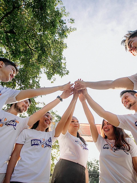 A group of people wearing matching T-shirts are standing in a circle and joining hands in the center, with trees and sky in the background.