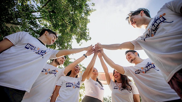 A group of people wearing matching T-shirts are standing in a circle and joining hands in the center, with trees and sky in the background.