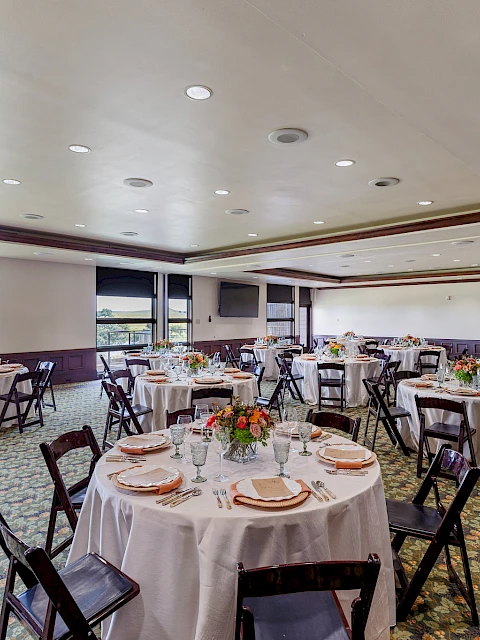The image shows a banquet room setup with round tables, white tablecloths, and floral centerpieces, with light streaming through large windows.
