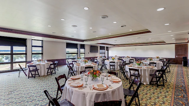 The image shows a banquet room setup with round tables, white tablecloths, and floral centerpieces, with light streaming through large windows.