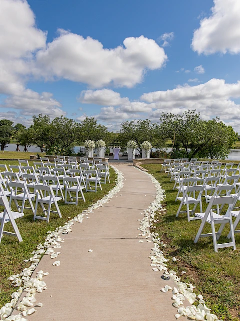 An outdoor wedding setup with white chairs, flower petals lining the aisle, and a sign, overlooking a scenic view with a lake and trees.