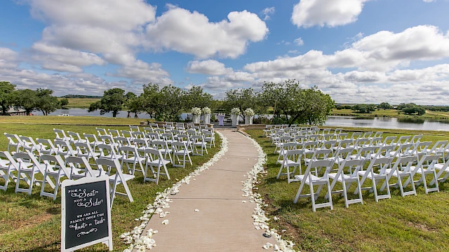 An outdoor wedding setup with white chairs, flower petals lining the aisle, and a sign, overlooking a scenic view with a lake and trees.