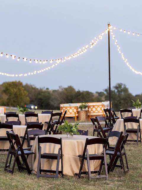 An outdoor event setup with tables and chairs on grass, draped in beige cloth, under string lights against a twilight sky.