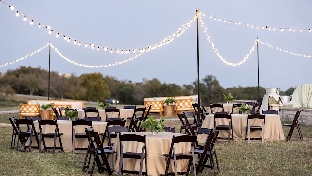 An outdoor event setup with tables and chairs on grass, draped in beige cloth, under string lights against a twilight sky.