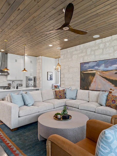 A modern living room with a beige sofa, colorful cushions, round table, and a kitchenette. Wooden ceiling and decorative elements adorn the space.
