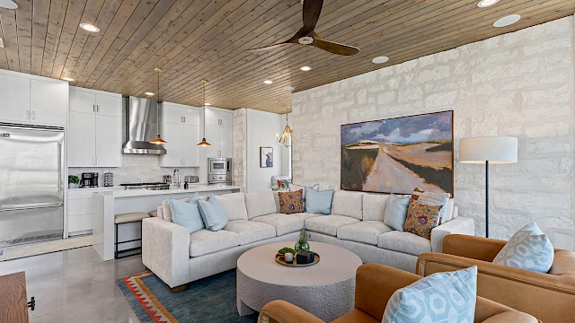 A modern living room with a beige sofa, colorful cushions, round table, and a kitchenette. Wooden ceiling and decorative elements adorn the space.