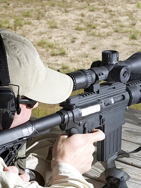 A person aiming a rifle with a scope at a shooting range, wearing ear protection and a cap, positioned on a wooden surface outdoors.