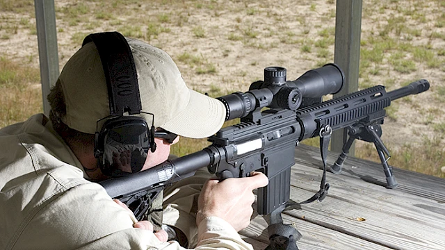 A person aiming a rifle with a scope at a shooting range, wearing ear protection and a cap, positioned on a wooden surface outdoors.