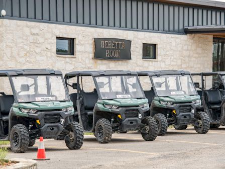 A row of four green all-terrain vehicles parked outside a stone building with a "Benefits BOW" sign.