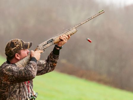 A person in camouflage attire aims a shotgun while ejecting a shell outdoors.
