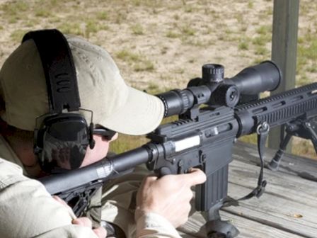 A person is aiming a rifle with a scope, wearing ear protection and a cap at a shooting range.