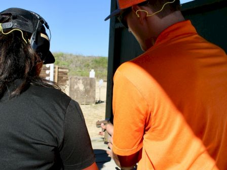 Two people wearing protective eyewear and caps are standing outdoors, possibly at a shooting range.
