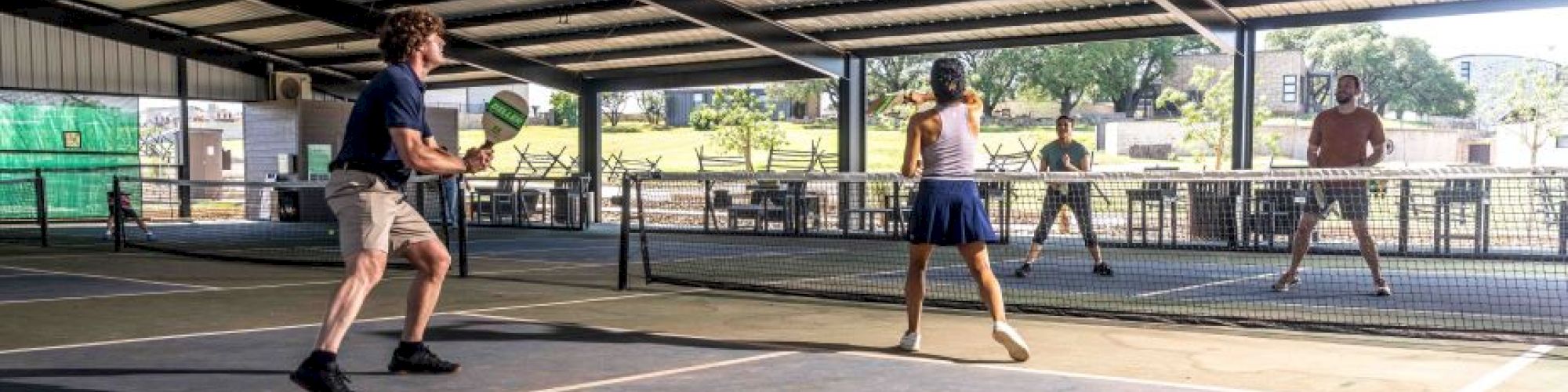 Four people are playing pickleball on an indoor court, with nets dividing the space, under a metal roof with natural light coming in.