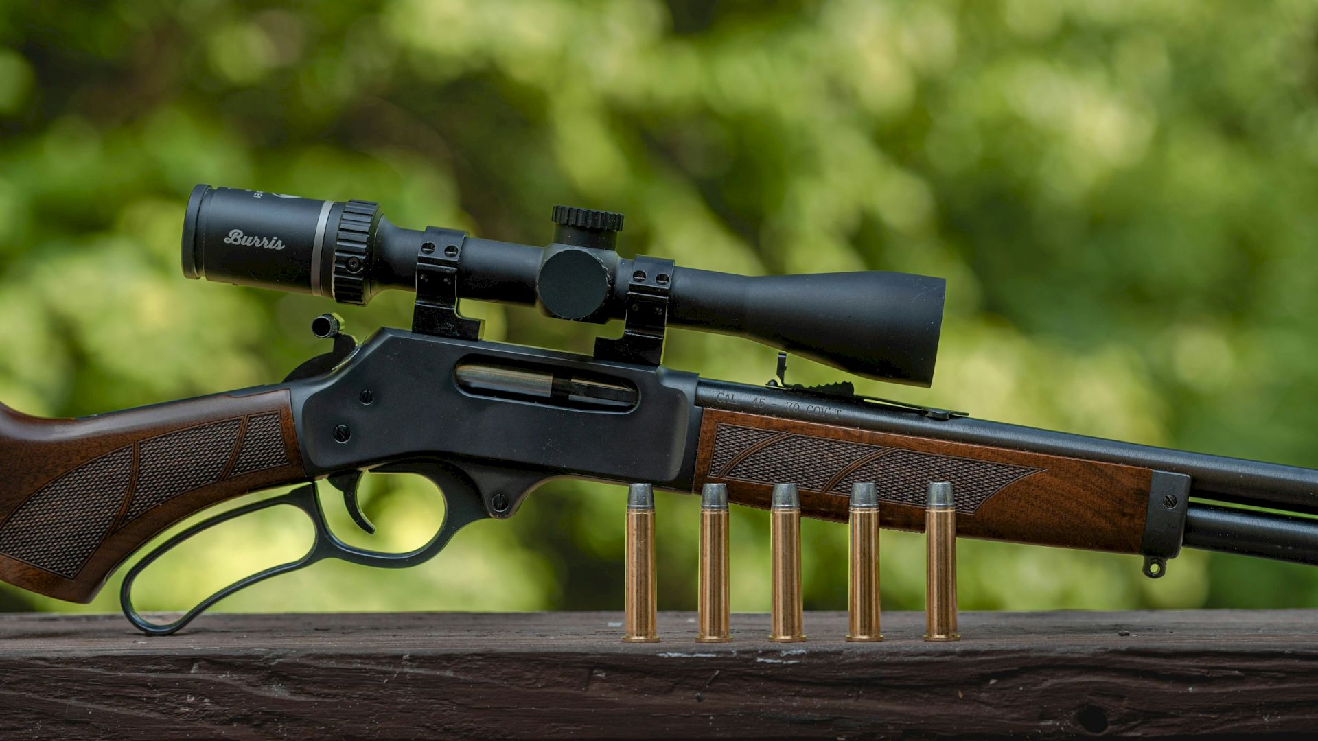 A scoped hunting rifle on a table with several brass cartridges, outdoors with a blurred green background.