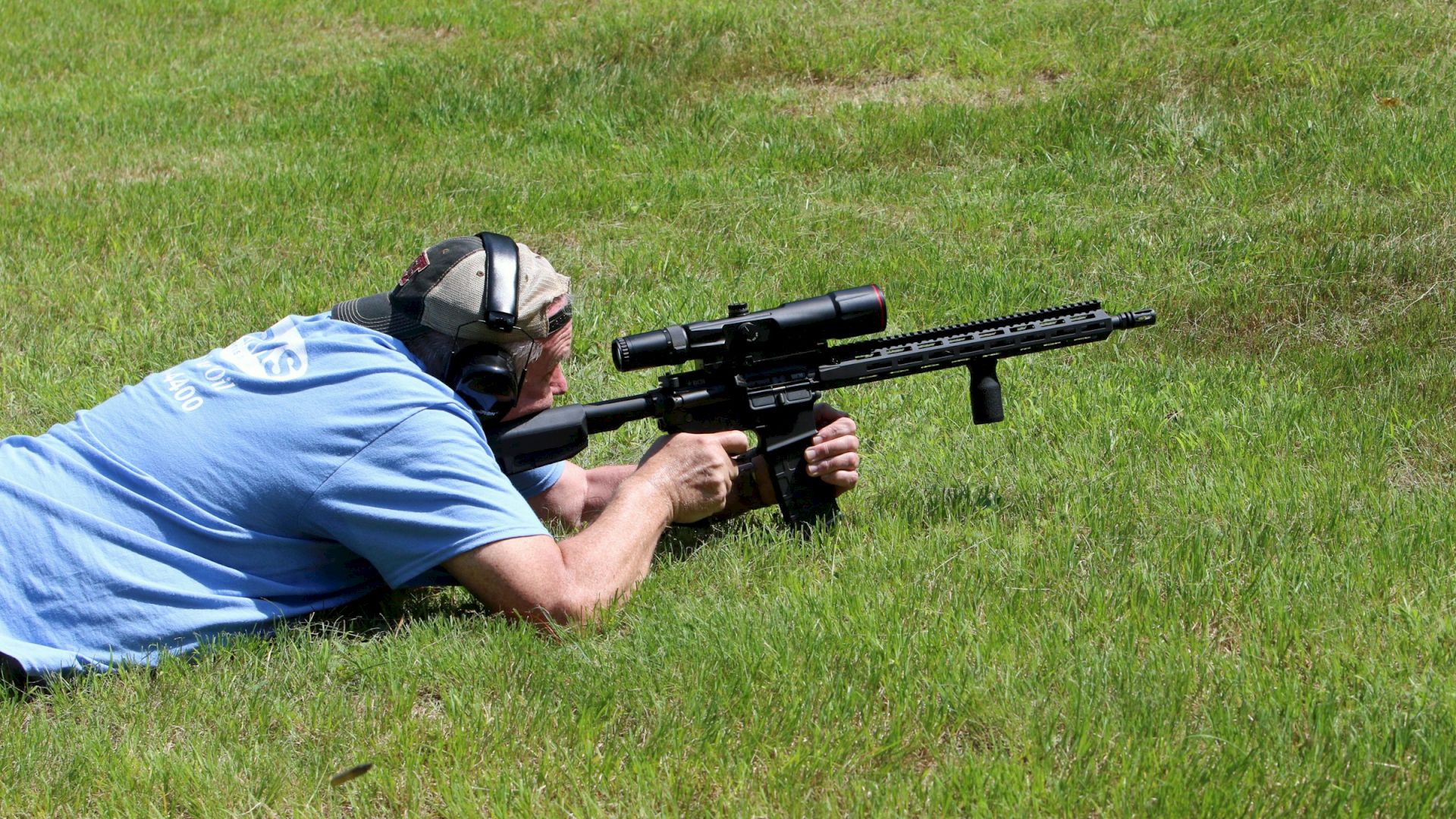 A person lying on grass, aiming a scoped rifle while wearing earmuffs for protection.