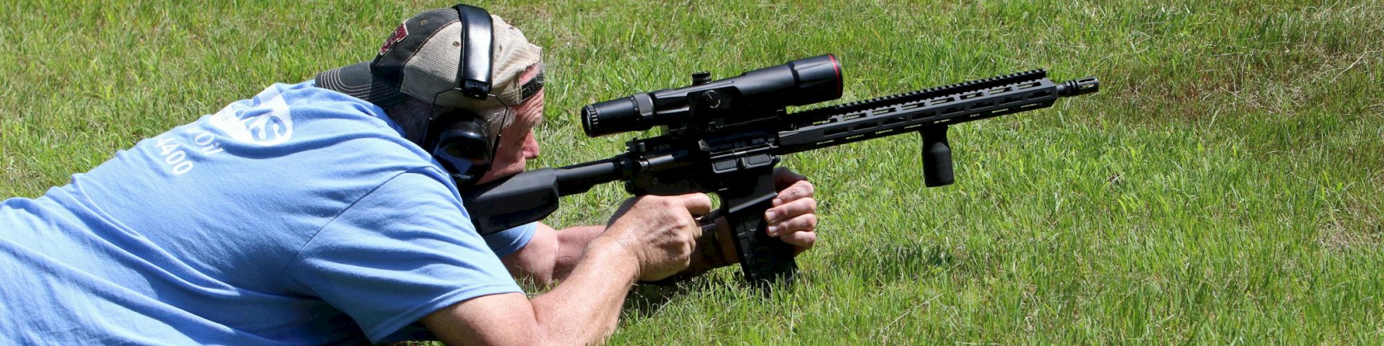 A person lying on grass, aiming a scoped rifle while wearing earmuffs for protection.