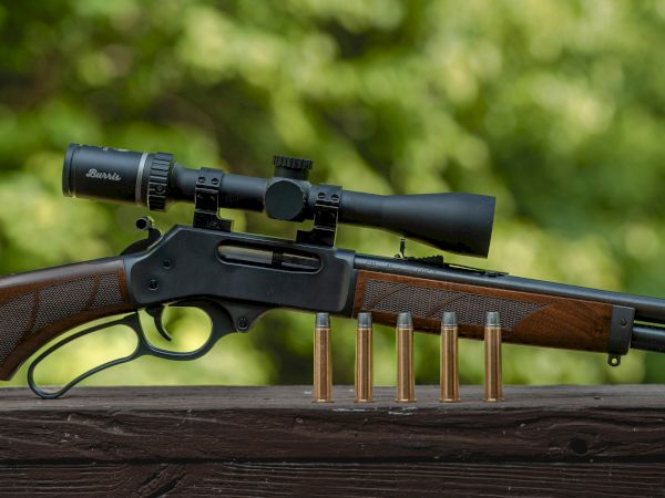 A rifle with a scope is resting on a wooden surface alongside five bullets, with a blurred foliage background.