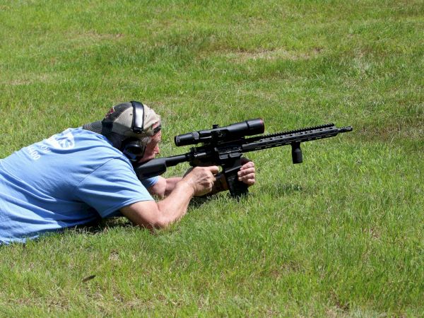 A person lies in the grass aiming a rifle with a scope, wearing a blue shirt and protective ear gear.