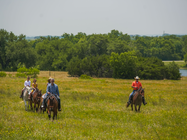 A group of people is horseback riding through a grassy field with trees and a small body of water in the background.