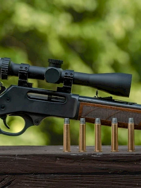 A rifle with a scope is displayed on a wooden surface, with five bullets standing upright in front of it.