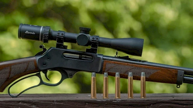 A rifle with a scope is displayed on a wooden surface, with five bullets standing upright in front of it.