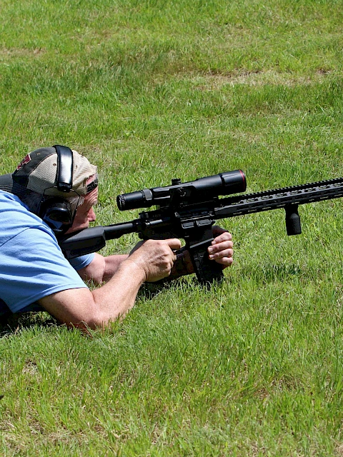 A person lying on grass aims a rifle with a scope, wearing ear protection, in a shooting position.