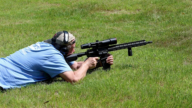 A person lying on grass aims a rifle with a scope, wearing ear protection, in a shooting position.