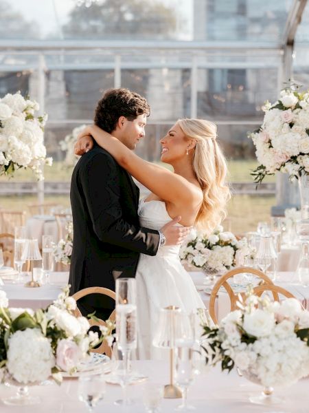 A couple is embracing in a beautifully decorated venue with white flowers and elegant table settings, suggesting a wedding celebration.