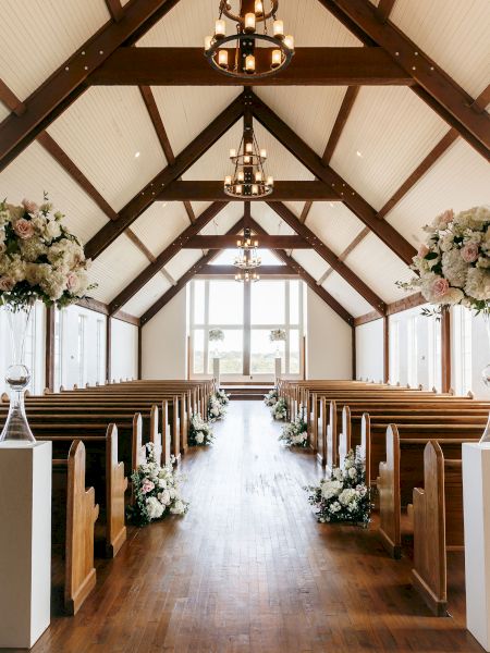 An elegantly decorated chapel interior with wooden pews and floral arrangements, lit by chandeliers, leading to a windowed altar area.