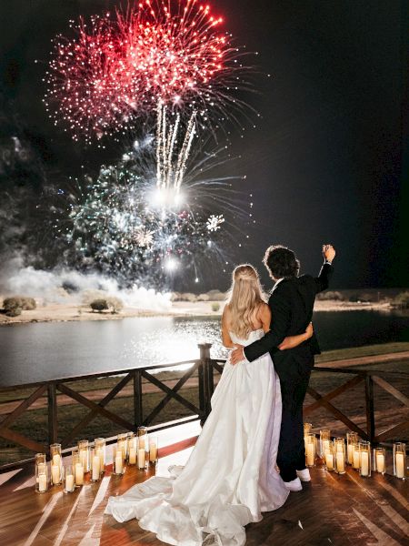 A couple embraces, watching a vivid fireworks display over a lake, surrounded by candles on a wooden deck at night.
