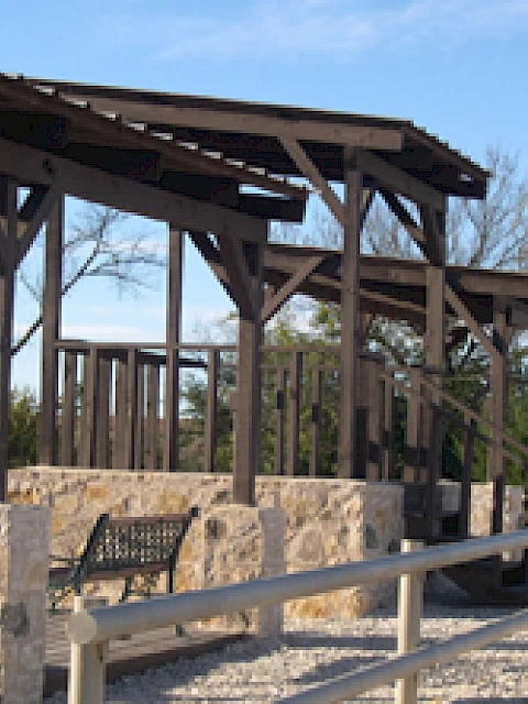 The image shows a shaded outdoor seating area with benches and stone accents, surrounded by trees, under a clear sky.