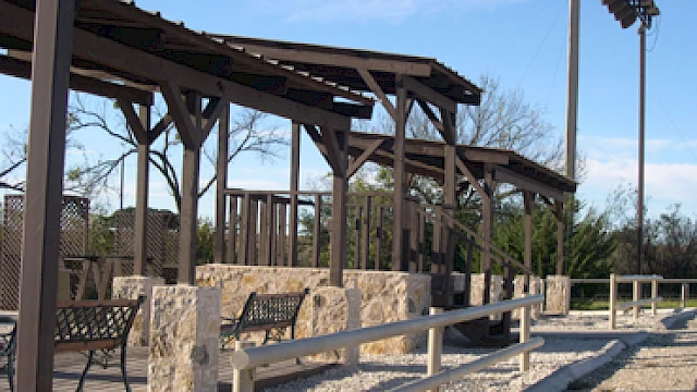 The image shows a shaded outdoor seating area with benches and stone accents, surrounded by trees, under a clear sky.
