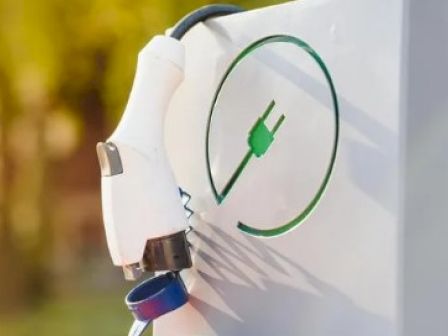A wall-mounted electric vehicle charger with a green plug icon and a white safety clip, ready for charging outdoors.