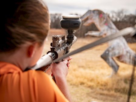 Person aiming a rifle with a scope at a target in an outdoor range; a target shaped like a dragon is visible in the distance.