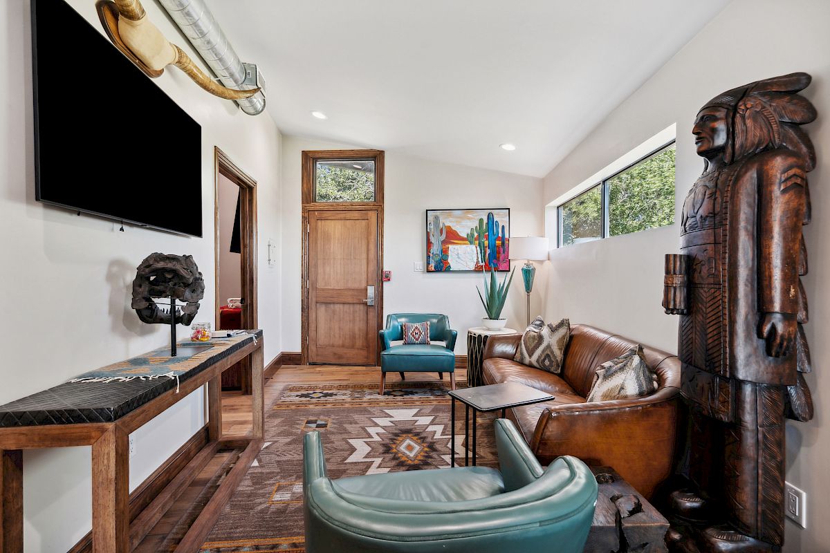 A stylish living room with a mounted TV, wooden accents, a large carved statue, leather couches, a teal chair, tribal rug, and bold wall art, bright and welcoming.
