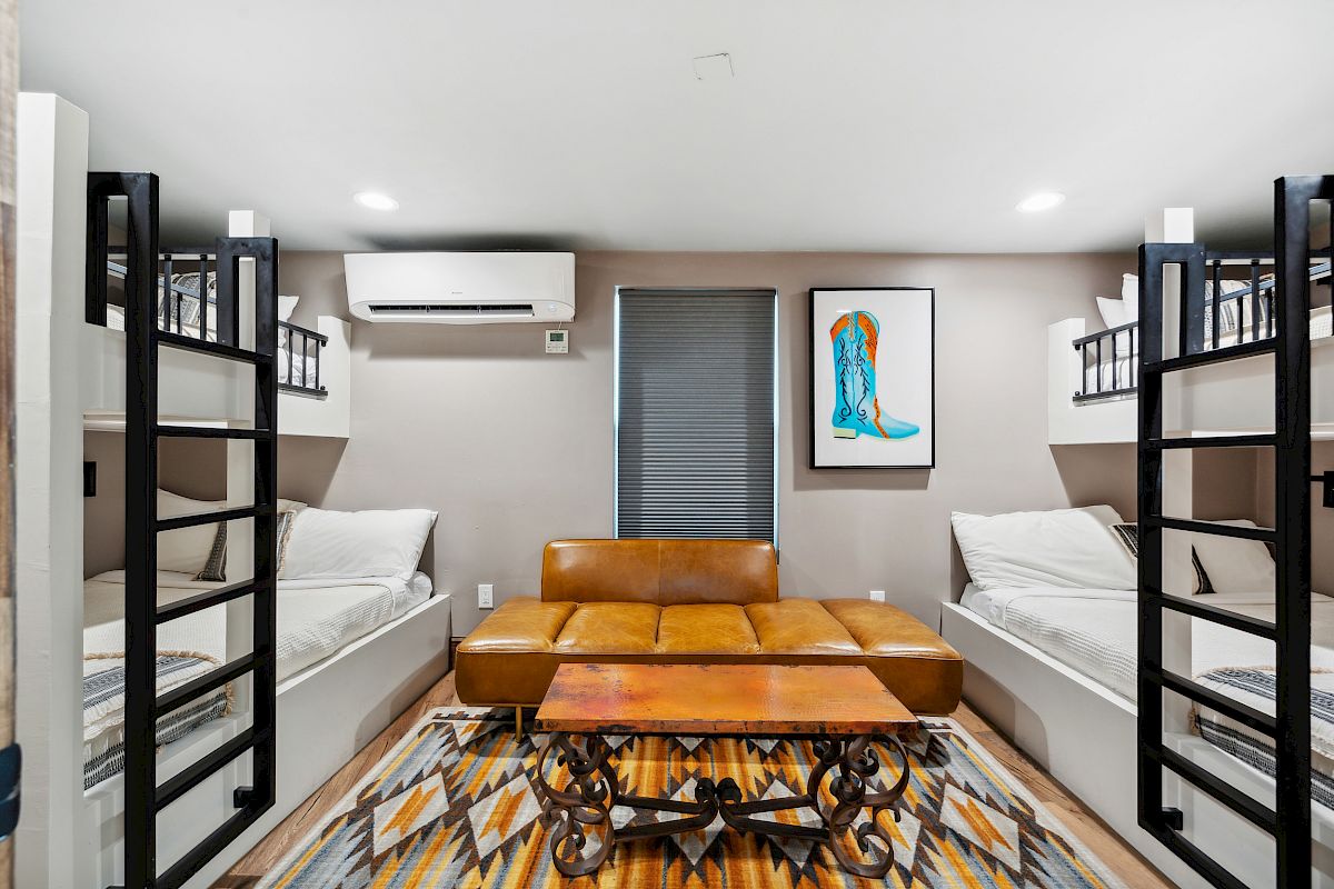 A compact dorm room with two bunk sets facing a central orange leather sofa and coffee table, framed by a patterned rug and wall art.